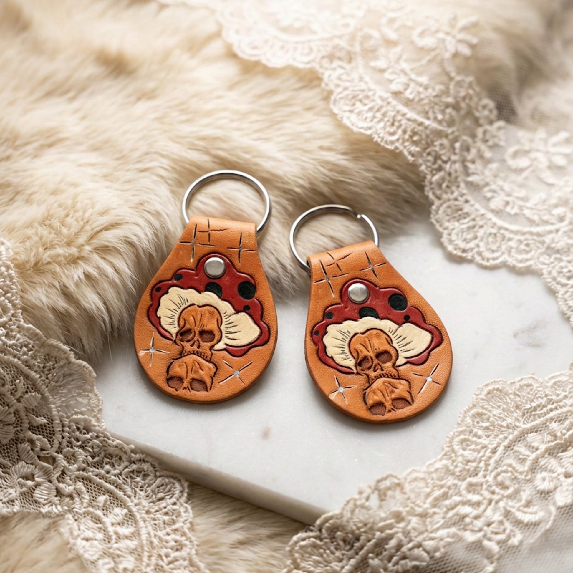Two hand-tooled tan leather keychains with a skull and red mushroom motif, styled on a fur and lace backdrop with a marble surface