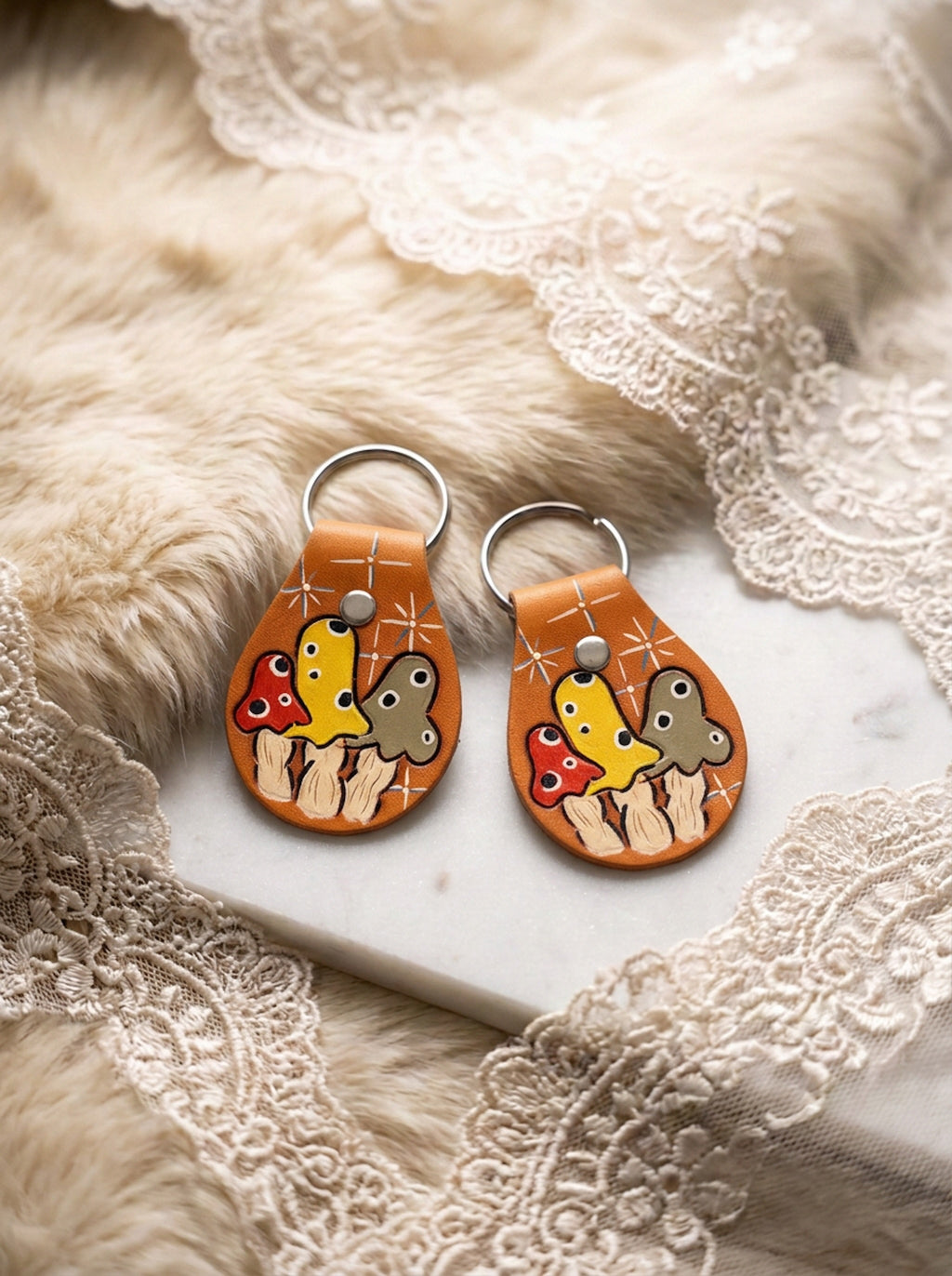 Two hand-tooled warm tan leather keychains with a colorful mushroom trio motif in red, yellow, and taupe, styled on a fur and lace backdrop