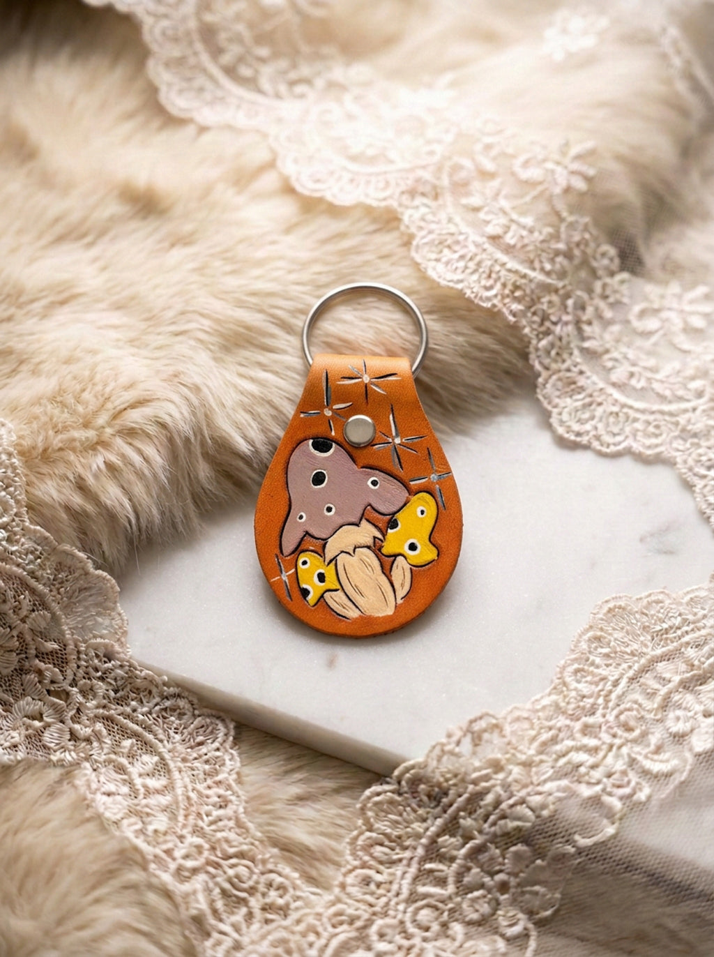 Single hand-tooled amber leather keychain with a wild mushroom cluster motif in taupe, yellow, and cream with etched sparkle details, on a fur and lace backdrop
