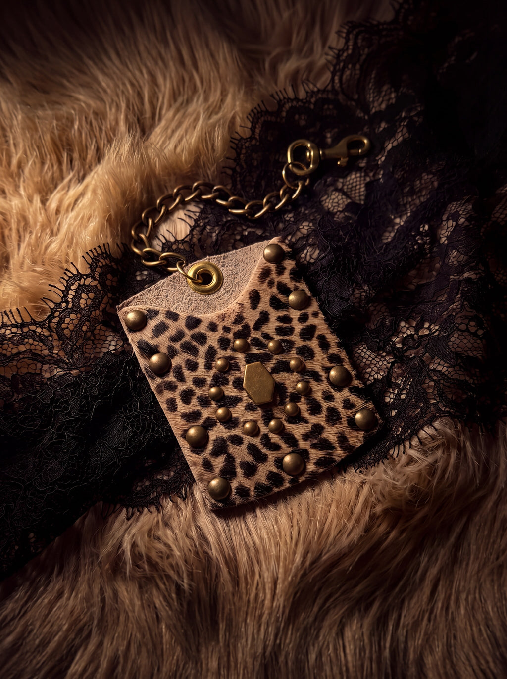Leopard print hair-on hide mini wallet with antique brass dome studs, hexagon snap closure, and brass chain, styled on a fur and black lace backdrop